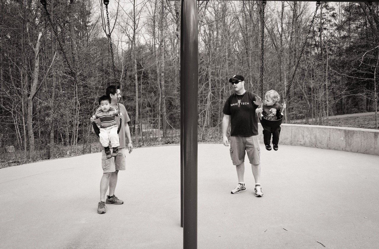 monochrome image of two dads pushing their children on swings. A bar bisects the image