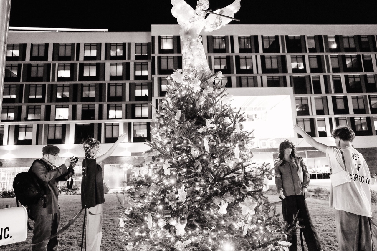 a black and white image of a young photographer looking similar to the mannequins dummies standing by a Christmas tree 