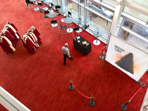 View from above of man walking toward a table and choir robes are on a rack. A selfie background says 'Christmas at DPAC'