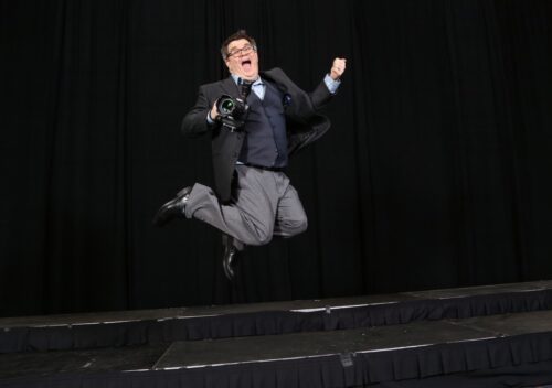 Ken Huth in a suit and with a camera in-hand, leaps into the air for joy and looks a bit silly
