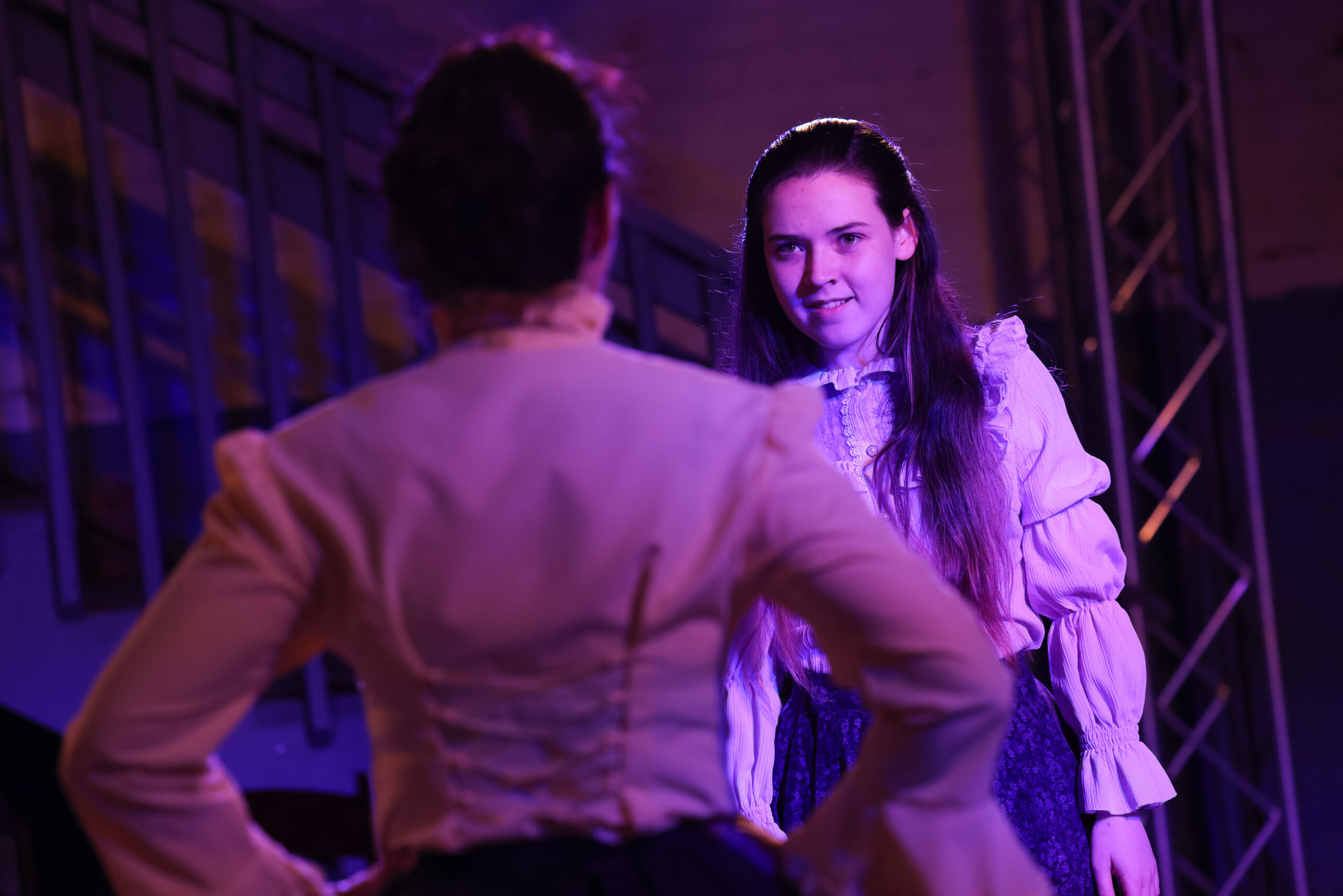 magenta theatre lighting with a young female actor glaring at another actor with her back to the camera