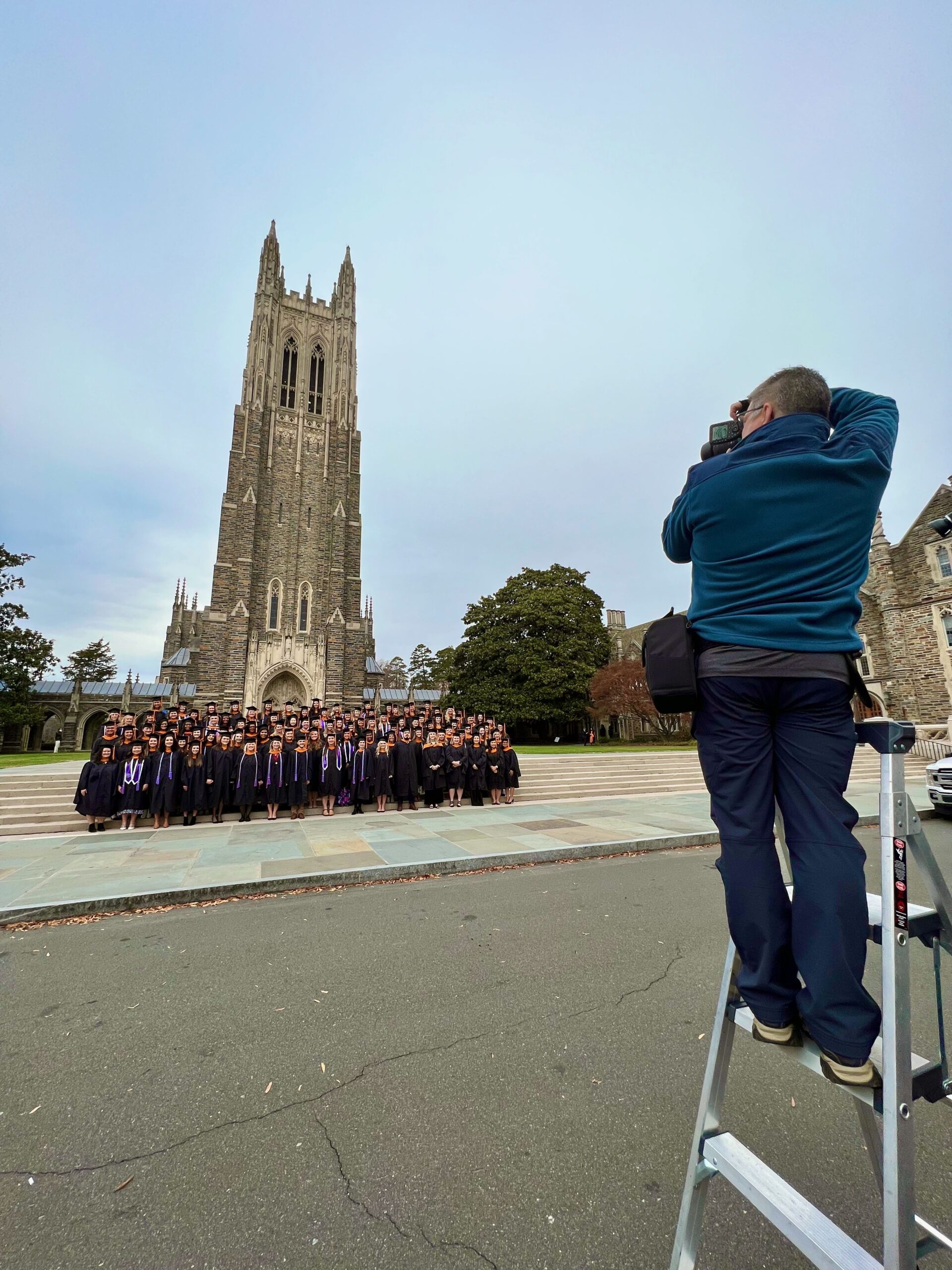photographer Ken Huth stands on top of a tall ladder to photographer a large group of graduates in front of Duke Chapel