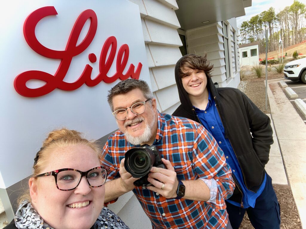 with red Eli Lilly sign behind them Ken Huth in plaid shirt and holding camera is with young assistant Oliver Huth and another smiling female employee