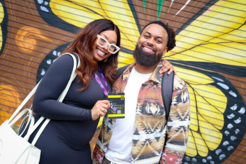 Two people stand in front of a butterfly mural in downtown Durham holding badges for the Griot and Grey Owl Conference.