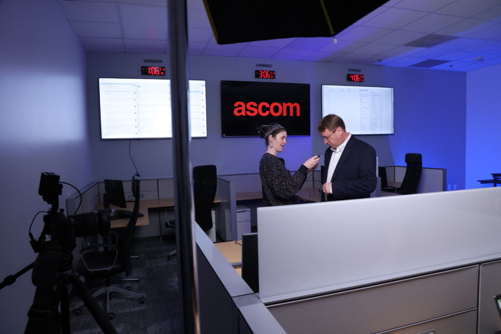HuthPhoto Team member Erin attaches a small microphone onto a businessman's suit coat in a blue-lit room with the company name ASCOM on one of three large video screens