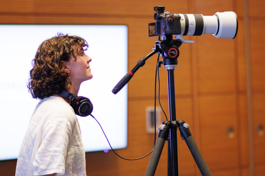 Videographer Erin Scannell with a camera and large lens. She has headphones around her neck and the camera is on a very tall tripod