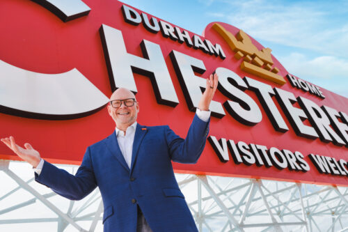 portrait of Geoff Durham with the large red Chesterfield "Durham Visitors Welcome" sign
