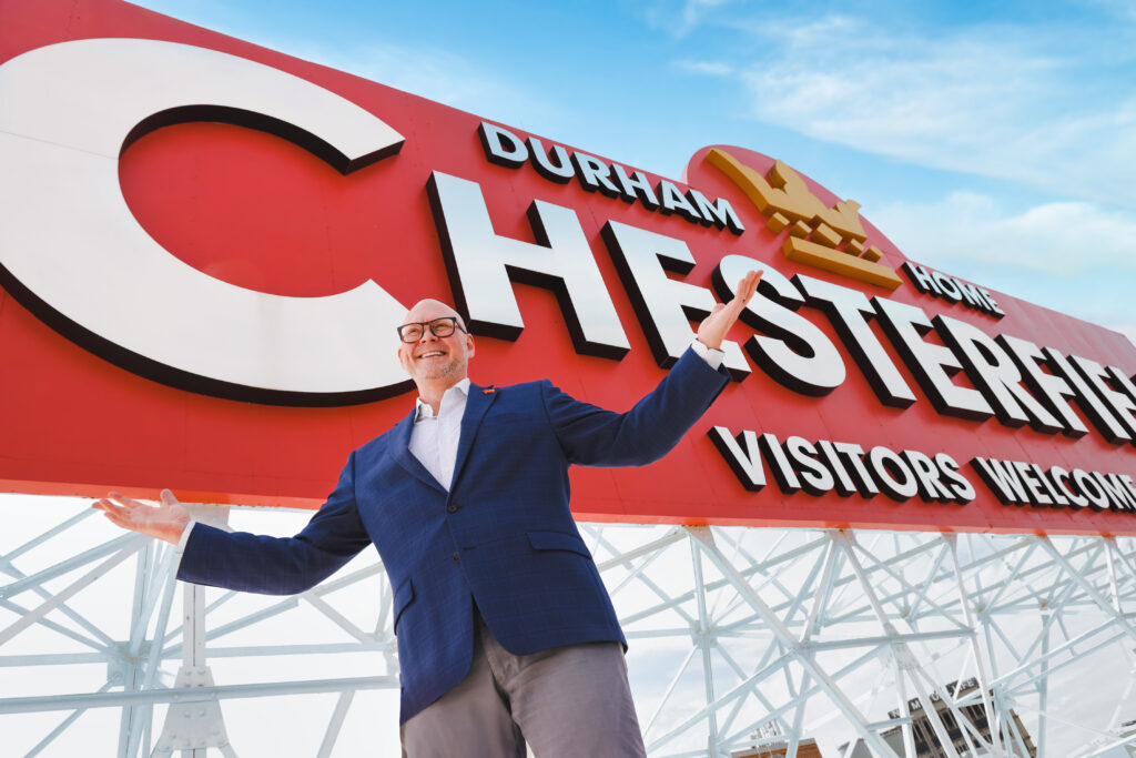 portrait of Geoff Durham with the large red Chesterfield "Durham Visitors Welcome" sign