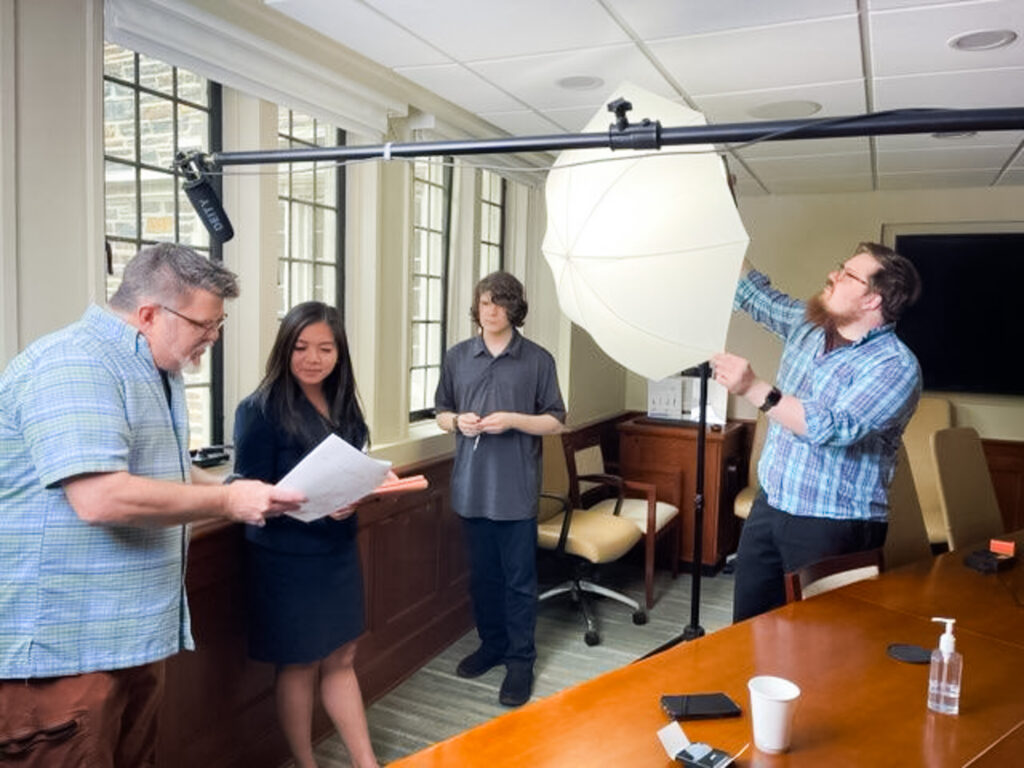 Ken Huth reviews a script with a video subject as young assistant Oliver Huth and bearded Colin Huth adjusts a large photo light with umbrella