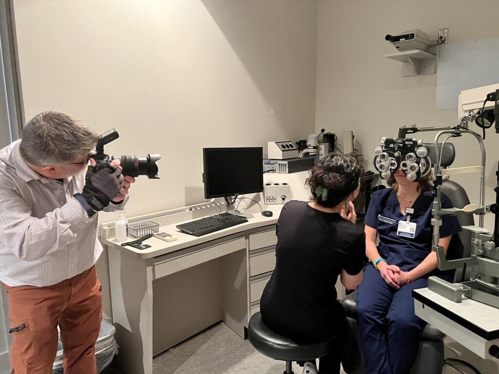 photographer Ken Huth on left of image holds a large camera photographing young eye tech trainees use equipment on the right