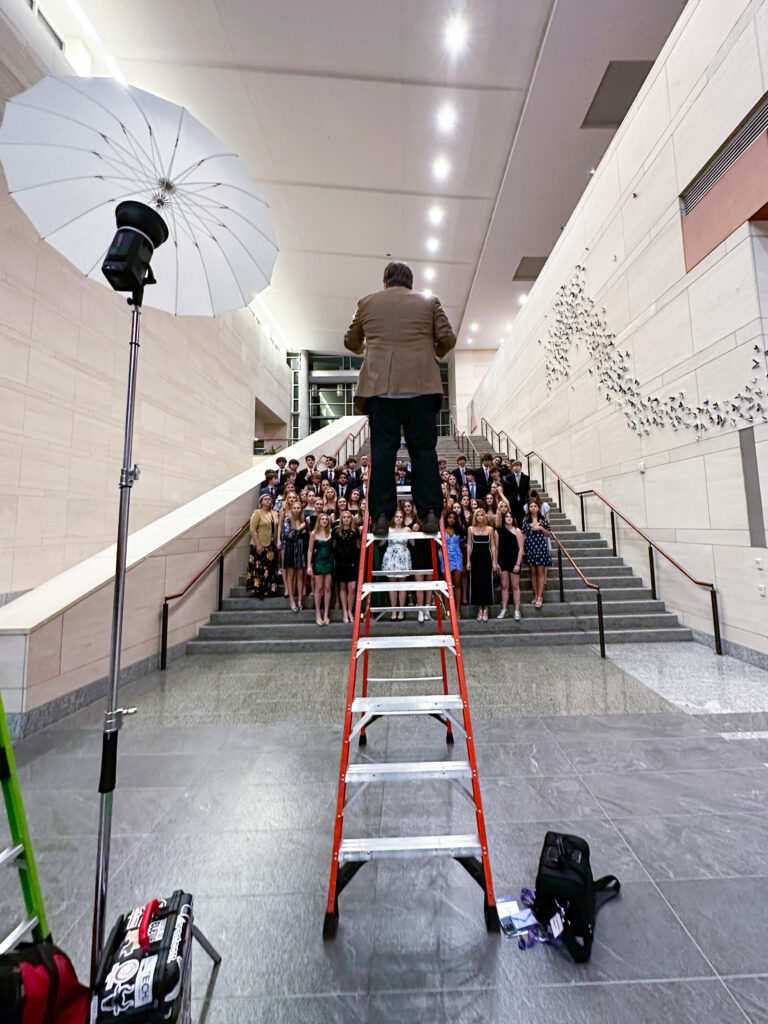 Photographer Colin Huth from behind is up a VERY large ladder with a photo light and umbrella on a stand nearby. He is photographing a large group of high school student on stairs at the Raleigh Convention Center