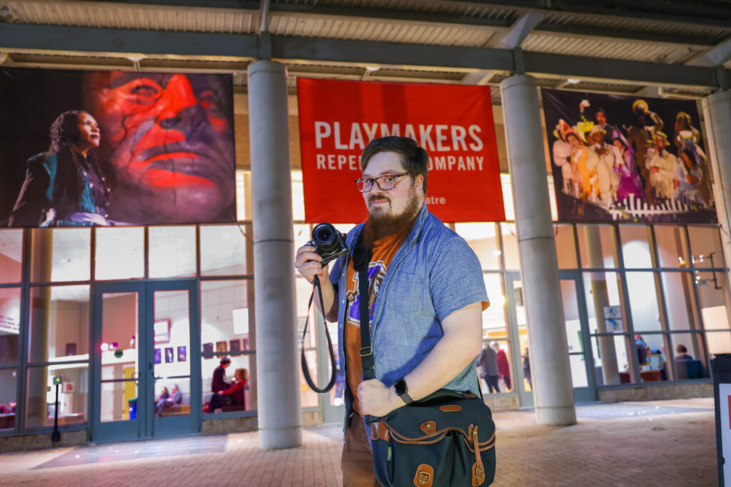 Young photographer Colin Huth smiles at the camera while holding a small camera. Behind him are banners and images he has taken for PlayMakers Rep Theatre