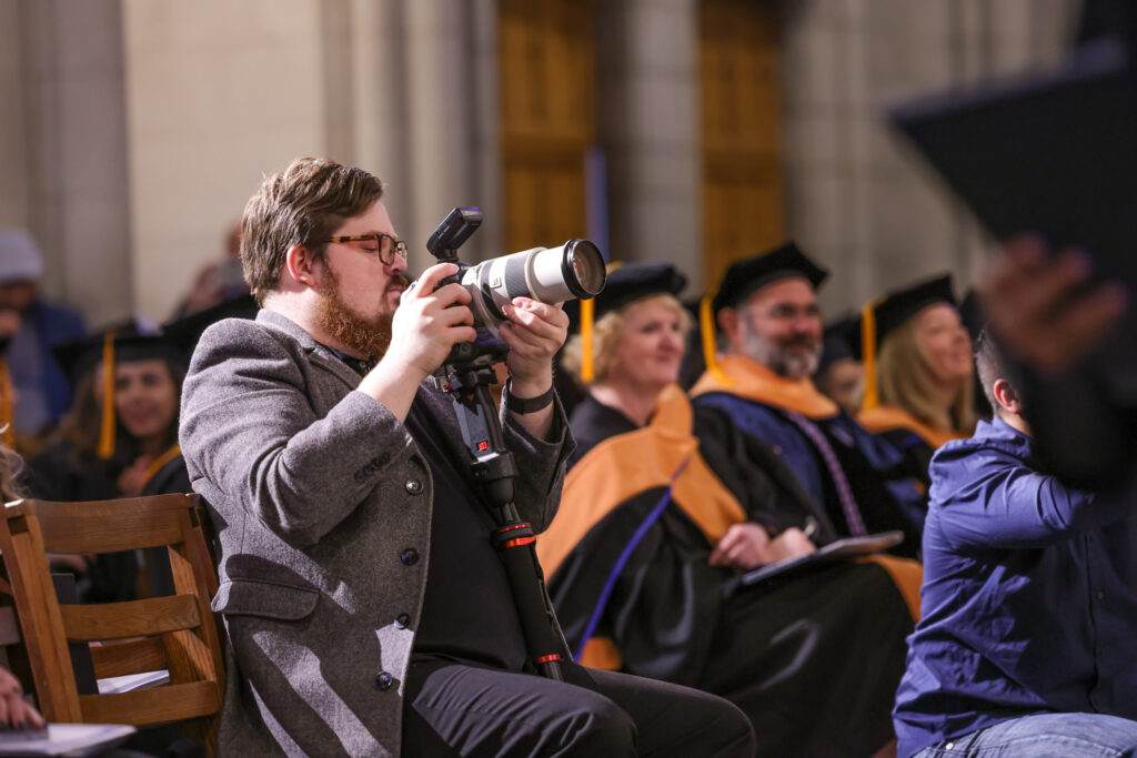 Photographer Colin Huth in a suit coat with a long camera lens photographs graduates from a seat in the audience at Duke Chapel
