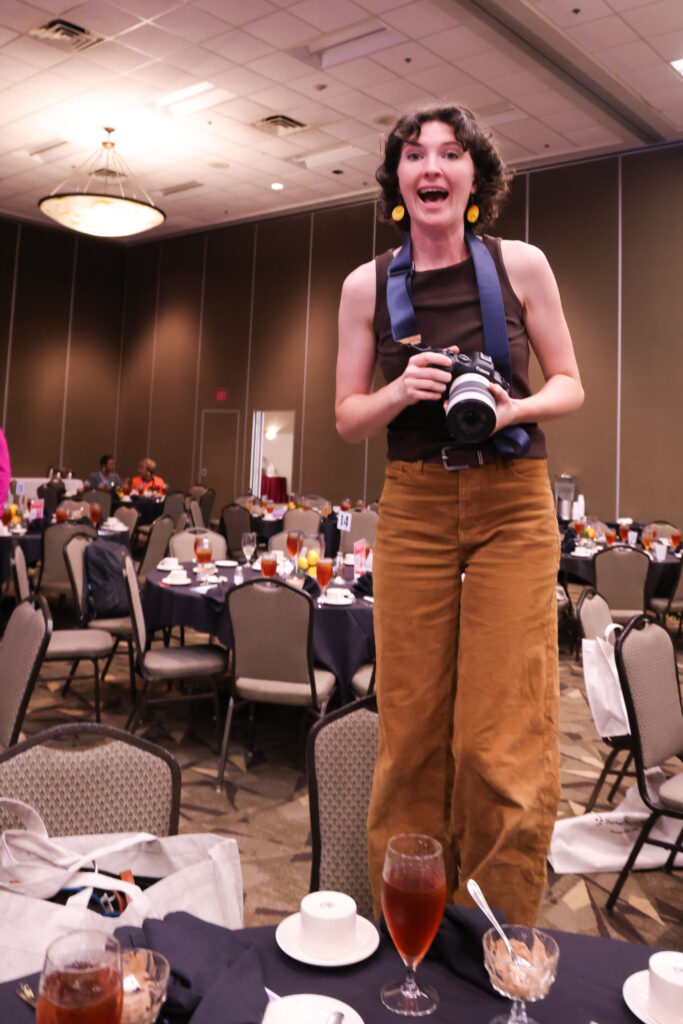 Photographer Erin Scannell stands on top of a chair to get a photo and laughs at having her photo taken