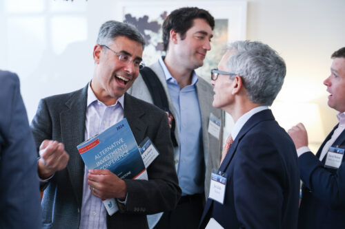 business man speaks with a big smile to others at a business conference. He's holding a program for a conference that says "Alternative Investing Conference"