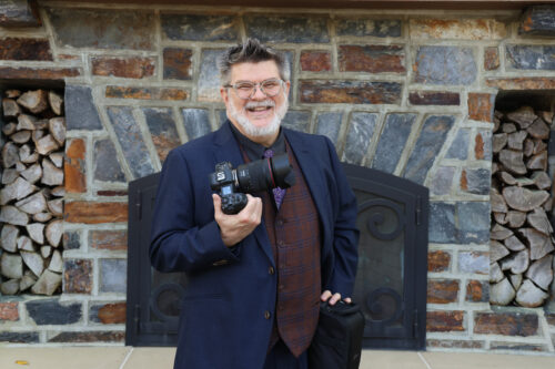 man standing looking at the camera with lovely bricks and log fireplace behind him
