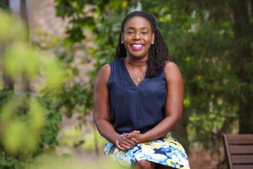 image of a young black woman in a blue top and skirt smiling at the camera surrounded by green leaves outdoors