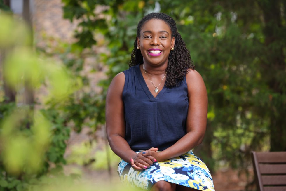 image of a young black woman in a blue top and skirt smiling at the camera surrounded by green leaves outdoors