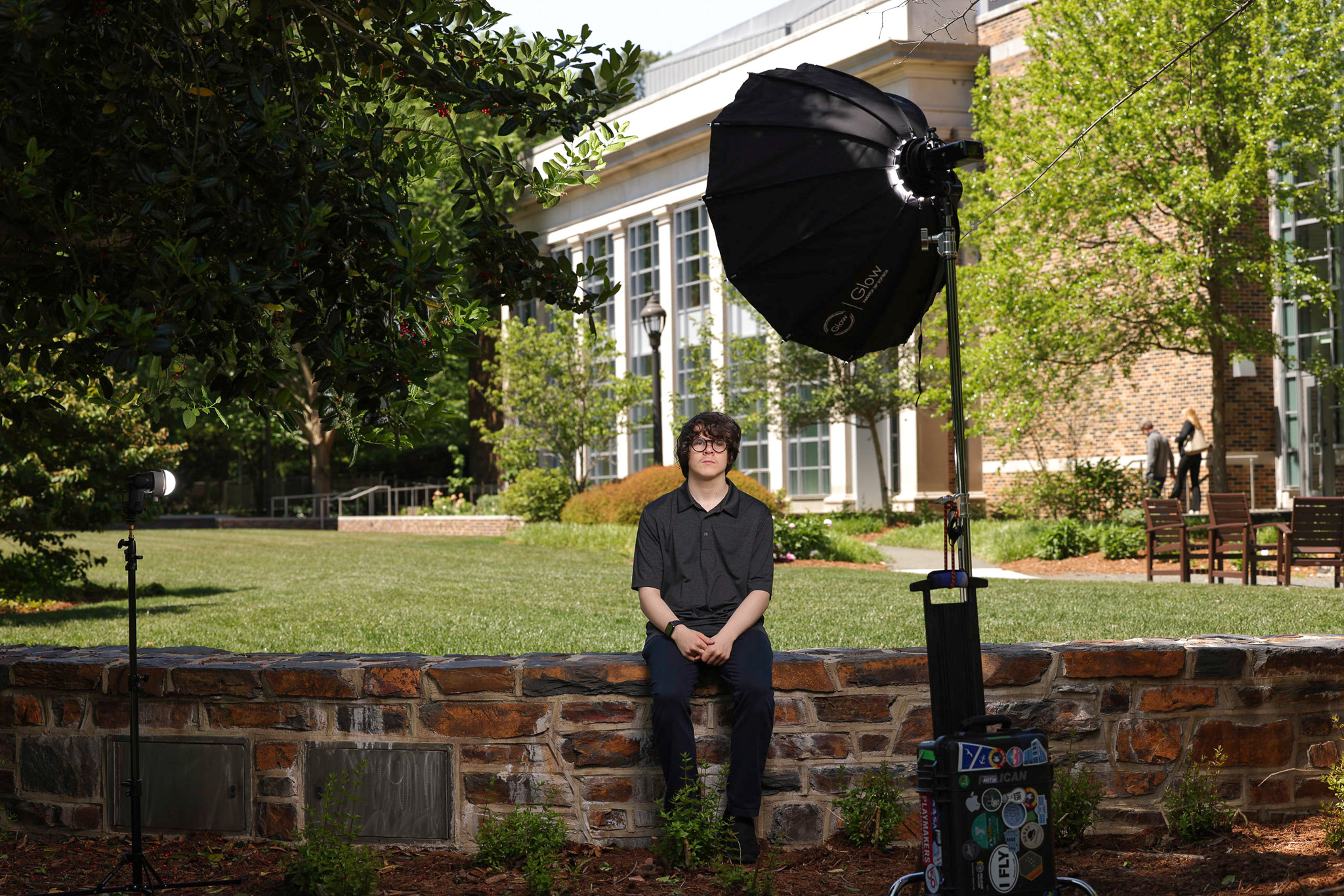 young man sits on a wall with photo lights nearby. Background is an Ivy League looking building (Duke Law)