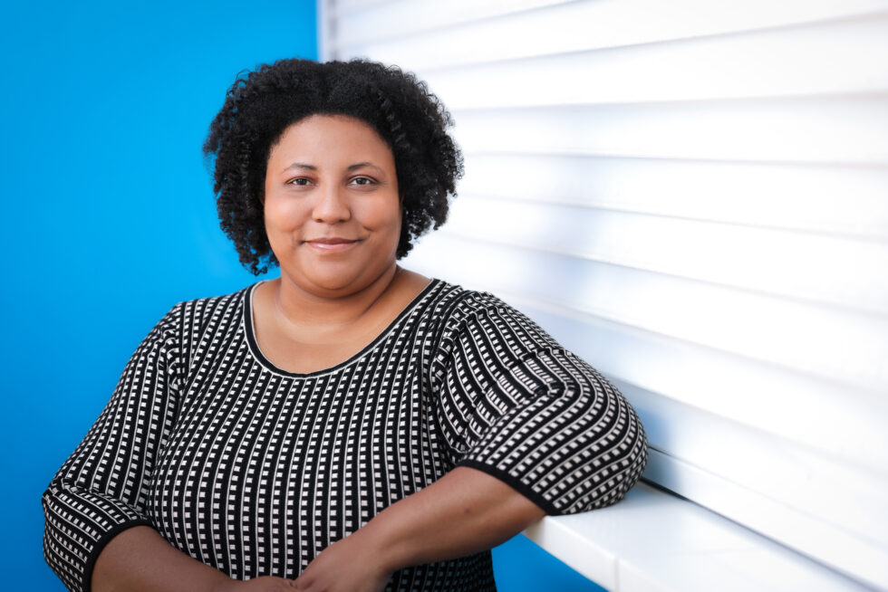 image of a black woman with short hair leaning on a counter edge. The background has a strong blue and white design
